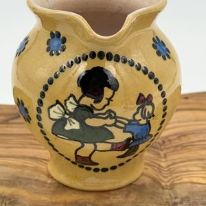May include: A yellow ceramic jug featuring a hand-painted illustration of two children dancing inside a dotted black circle. The jug has a glossy finish, a wide spout, and additional blue flower motifs painted around the upper body.