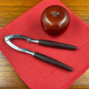 May include: A silver nutcracker with dark wood handles sits on a red linen napkin with a wooden apple beside it.