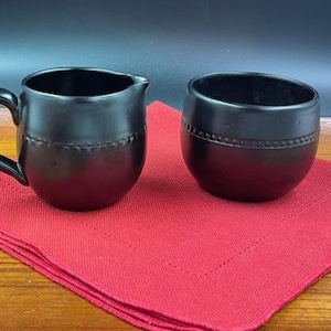 以下が含まれることがあります： 黒色のセラミック製のクリーマーとシュガーボウルセット。各ピースの中央にテクスチャ加工されたバンドが付いています。セットは赤いナプキンの上に置かれています。