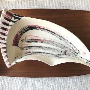 Puede incluir: Una fuente de cerámica color crema, con una forma curva única, sobre una bandeja rectangular de madera. El plato presenta pinceladas negras y granates y un borde a rayas. La bandeja de madera es de un marrón oscuro intenso.