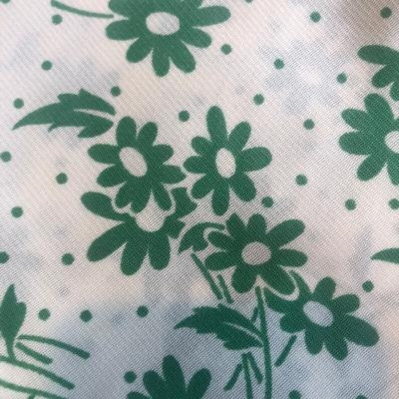 May include: White fabric with a repeating pattern of green daisy flowers. The daisies have white centres and are arranged in a cluster.