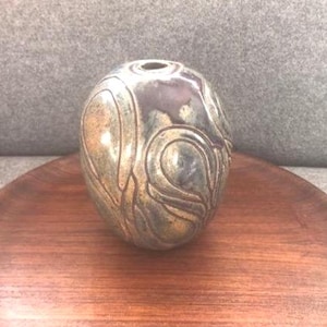 May include: A small, round ceramic vase with a swirling, abstract design in shades of brown, green, and gold.