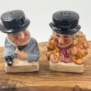 May include: Two ceramic salt and pepper shakers shaped like figures wearing black top hats. One figure wears a blue coat and the other wears a yellow coat. Both figures have painted facial features and are set on rectangular bases with a light tan finish.