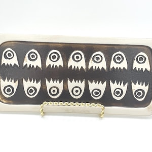 Könnte beinhalten: Rechteckige Keramikschale mit dunkelbrauner Mitte und cremefarbenem Rand. Die Mitte zeigt ein sich wiederholendes Muster aus weißen geisterartigen Figuren und blütenartigen Formen. Die Schale wird auf einem goldfarbenen Metallständer präsentiert.