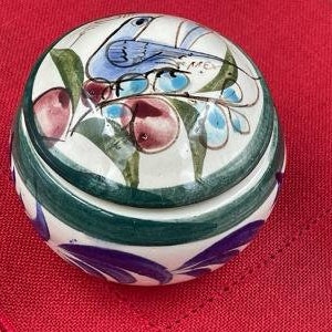 May include: A round, ceramic trinket box with a lid. The box is white with a hand-painted design featuring a blue bird, green leaves, and red and blue floral accents. The box has a green and purple striped border.