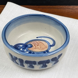 以下が含まれることがあります： 青い縁取りと青と茶色の猫のデザインが施された白い陶器のボウル。ボウルには底に「KITTY」と書かれています。