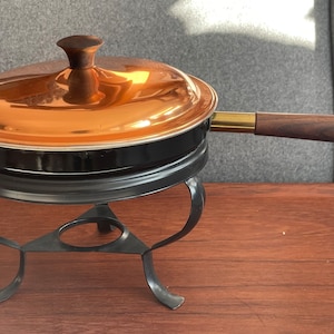 Puede incluir: Una olla negra con tapa de cobre y mango de madera, sobre un soporte de metal negro. La olla tiene un borde dorado y un pomo de madera oscura en la tapa. El soporte tiene un diseño curvo decorativo.