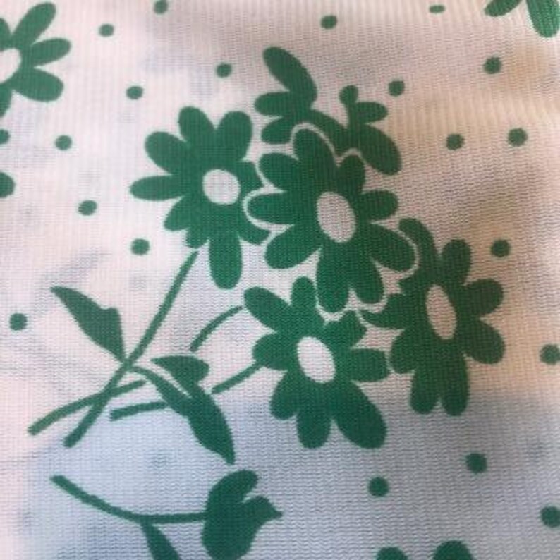 May include: White fabric with a green floral pattern. The flowers are stylised daisies with white centres and green petals. The fabric has a subtle texture.