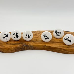 May include: Six white ceramic cabinet knobs with black text are displayed on a wooden board. The knobs feature the numbers 2, 3, and 4, and the phrases "no no no", "Private Keep Out", and "Junk". The board is made of wood.