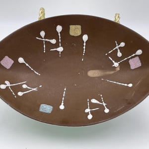 May include: Brown ceramic serving dish with a unique, elongated oval shape. The dish is decorated with white, abstract designs and small, colorful square accents. The dish has two small handles and is supported by gold-colored metal stands.