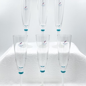 Puede incluir: Seis copas de champán transparentes con un acento azul en la base del tallo. Cada copa tiene un logotipo impreso en la parte delantera. Las copas están dispuestas en forma de pirámide sobre una superficie blanca.