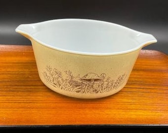 May include: A white ceramic casserole dish with a brown mushroom and leaf design. The dish has two handles and is on a wooden surface.