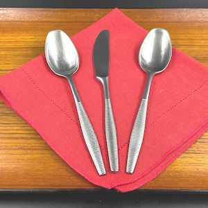 May include: A stainless steel knife and two spoons are arranged on a red napkin, set on a wooden tray. The cutlery has a ribbed handle design. The napkin is square and has a stitched border.