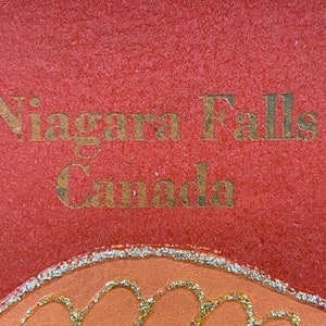 May include: A red background with gold lettering that reads "Niagara Falls Canada".
