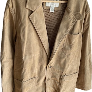 May include: A tan suede blazer with a notched lapel, two front pockets, and a button closure. The blazer has a classic design and is hanging on a wooden hanger. The interior has a mesh lining and a tag that reads "ORVIS".
