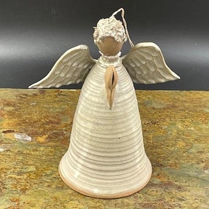 Puede incluir: Un adorno de ángel de cerámica blanca con un cuerpo en forma de campana y alas. El ángel tiene una superficie texturizada y un diseño simple.