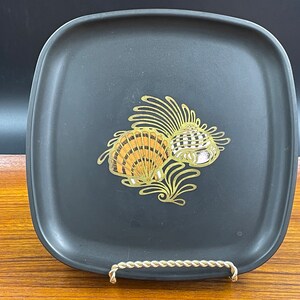 May include: A square, black serving tray with a gold seashell design. The seashells are detailed with orange and white accents. The tray is displayed on a gold metal stand.