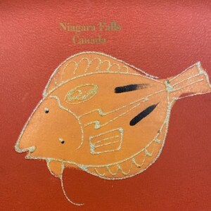 May include: Orange fish with black markings on a red background. The text "Niagara Falls Canada" is printed in gold lettering.