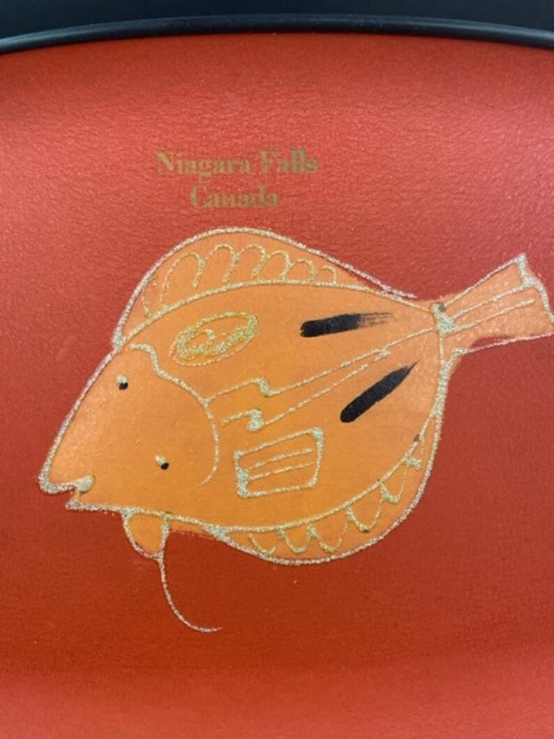 May include: Orange fish with black markings on a red background. The text "Niagara Falls Canada" is printed in gold lettering.