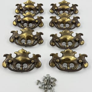 May include: Eight brass-toned metal drawer pulls with ornate, curved backplates and matching swing handles. Each pull features a decorative, symmetrical design with a dark, aged finish. A small pile of silver-coloured metal mounting screws is included.