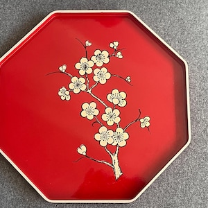 May include: Red octagonal tray with a white rim and a floral branch design. The branch features white blossoms with black detailing. The tray is likely made of lacquered wood or a similar material.