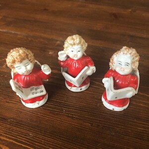 May include: Three porcelain figurines of angels singing from sheet music. The angels are wearing red robes with white trim and have blonde curly hair.