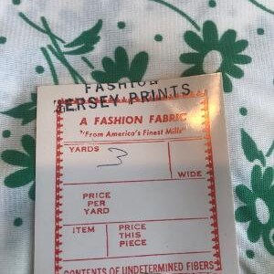 May include: A white fabric swatch with a green floral print. The swatch has a red border and text that reads "Fashion Jersey Prints, A Fashion Fabric, From America's Finest Mills, Yards, Wide, Price Per Yard, Item, Price This Piece, Contents of Undetermined Fibres, RN 34047".