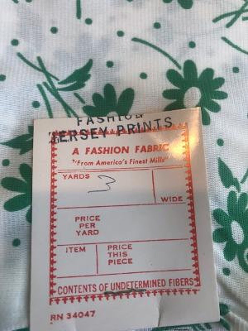 May include: A white fabric swatch with a green floral print. The swatch has a red border and text that reads "Fashion Jersey Prints, A Fashion Fabric, From America's Finest Mills, Yards, Wide, Price Per Yard, Item, Price This Piece, Contents of Undetermined Fibres, RN 34047".