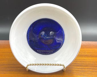May include: A round glass bowl featuring a wide white outer rim and a deep cobalt blue centre. The interior surface of the bowl displays a smooth, glossy finish with subtle swirling patterns within the blue glass, supported by a gold-toned stand.