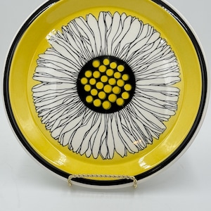May include: A ceramic plate with a large sunflower design. The plate features a yellow background with a black and white sunflower in the center. The sunflower has white petals and a black center with yellow dots. The rim is black and white.
