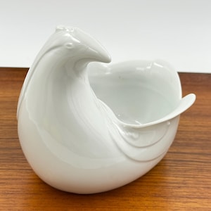 Puede incluir: Un cuenco decorativo de cerámica blanca con forma de pájaro. El cuenco tiene un diseño curvo con una boquilla en forma de pico y un cuerpo redondeado. Está sobre una superficie de madera.