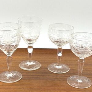 May include: Four clear, etched glass goblets with floral and leaf designs. The goblets have a slender stem and a round base, and are arranged on a wooden surface. The glasses are likely for serving wine or other beverages.