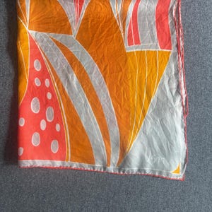 May include: A square scarf featuring a vibrant abstract pattern in shades of orange, coral, and white. The design includes large curved shapes, vertical lines, and a section with white circular spots. The edges are finished with a coral-colored hem.