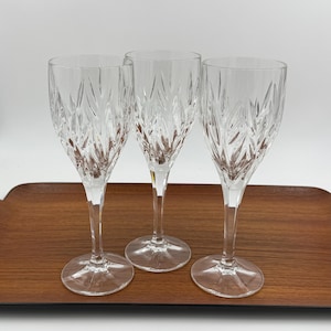 May include: Three clear crystal wine glasses with a detailed etched pattern, displayed on a wooden tray. The glasses have long stems and wide bowls, perfect for serving wine. The etched design adds a touch of elegance to the glassware.