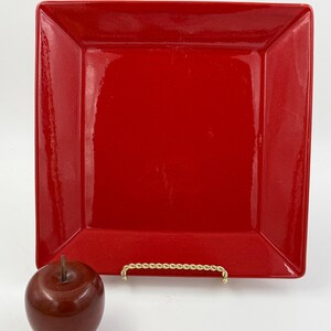 May include: A glossy, square, red plate with slightly raised edges. A decorative, brown apple sits to the left of the plate. The plate is displayed on a gold-colored, twisted-rope style stand.
