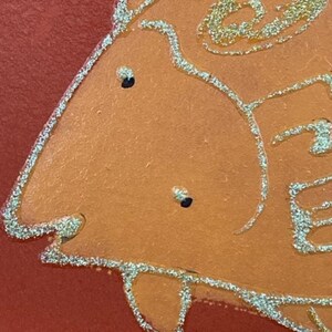 May include: Close-up of a stylized orange fish with gold glitter outlining the shape. The fish has two black eyes and a slightly open mouth.