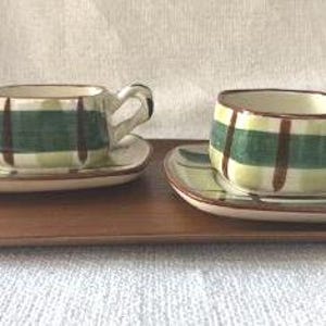 Puede incluir: Dos tazas de té y platillos de cerámica con un diseño de cuadros verdes y marrones, sobre una bandeja de madera. Las tazas tienen formas cuadradas y asas pequeñas. Los platillos son cuadrados con bordes redondeados.