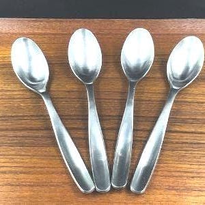 May include: Four stainless steel spoons arranged on a wooden surface. The spoons have a simple, elegant design with oval bowls and long, slender handles. The spoons are reflective, and the wooden surface has a warm, brown tone.