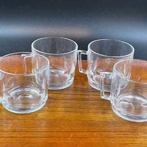May include: A set of four clear glass mugs with a smooth, cylindrical body and a square-shaped handle. Each mug features a thick, reinforced base and a simple, transparent design, arranged in a group to show their uniform shape and clear glass construction.
