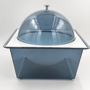 Puede incluir: Una cúpula de plástico transparente con un pomo plateado se asienta sobre una base cuadrada de plástico gris. La base tiene un borde blanco.