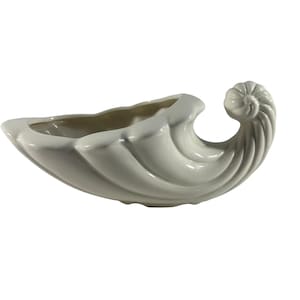 May include: A white ceramic seashell-shaped planter with a smooth, glossy finish. The planter has a curved, elongated form with a spiraled end and a central trough for holding plants or decorative items. The interior is a light beige color.
