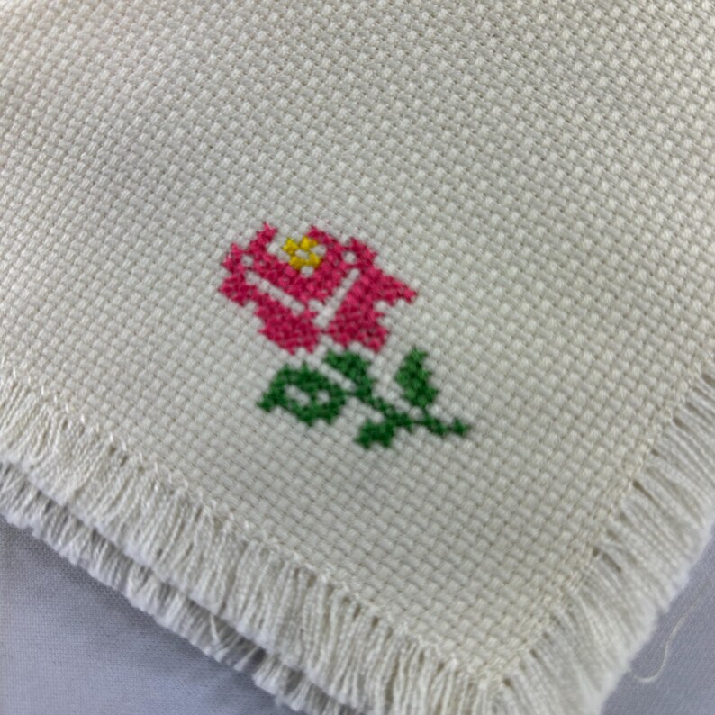 May include: A white fabric napkin with a pink and yellow embroidered rose design. The napkin has a fringed edge.