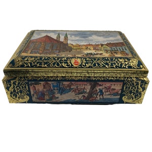 May include: A vintage blue and gold tin box with a detailed illustration of a European city scene. The lid features a cityscape with buildings and people, while the bottom depicts a horse-drawn carriage and figures in a rural setting.