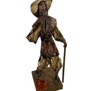 May include: A paper mache figurine of a person wearing a wide-brimmed hat and holding a walking stick. The figurine is standing on a wooden base with a small clay pot.