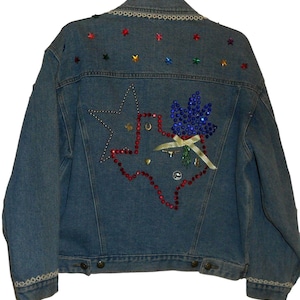 May include: A blue denim jacket with a large rhinestone design of the state of Texas on the back. The design includes a red, white, and blue star, a horseshoe, and a blue flower.