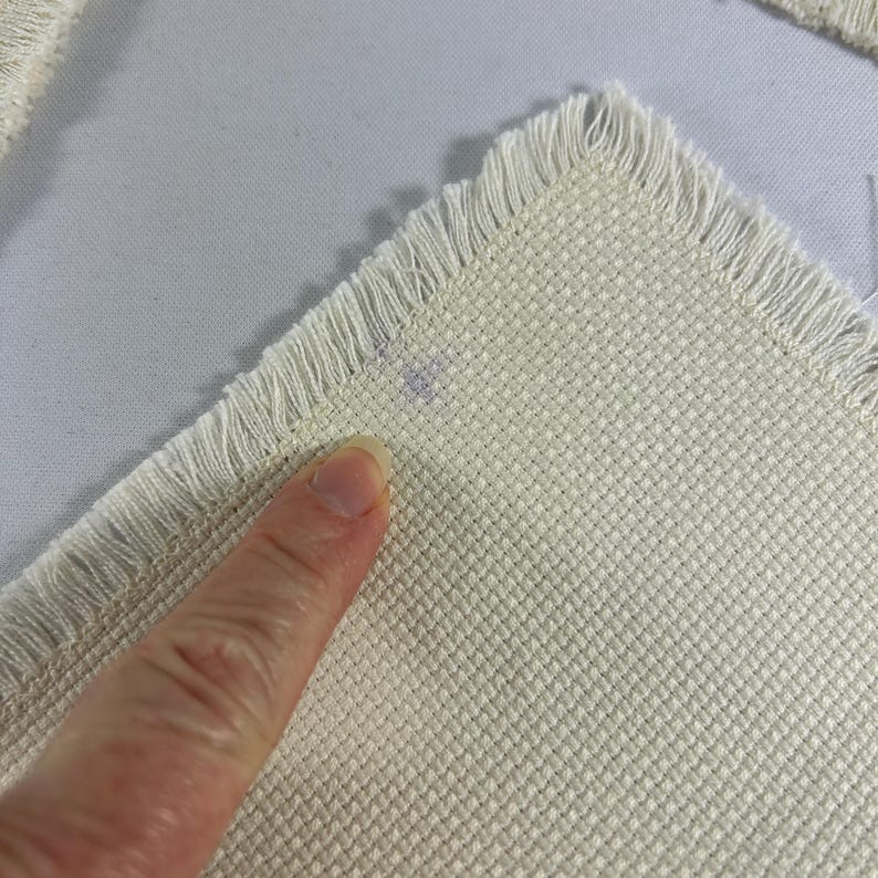May include: A white piece of Aida cloth with a fringed edge. The cloth is textured and has a small purple mark on the surface.