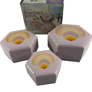 May include: Set of three lavender-colored, hexagonal boxes with seashell designs. The boxes have a yellow and tan fan-shaped seashell design on the lids. The boxes are labeled "Decorated Boxes" and are in a box that says "Set of 3 All Purpose".