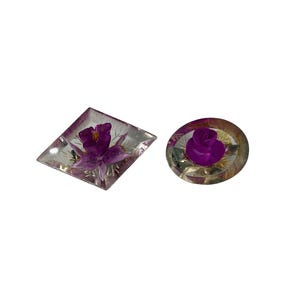 May include: Two resin paperweights. One is a clear, diamond-shaped paperweight with a purple flower. The other is a clear, round paperweight with a purple rose. Both have a floral theme.