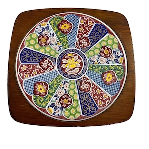 May include: A round ceramic tile with a colorful floral design in shades of red, blue, green, and yellow. The tile is set in a wooden frame.