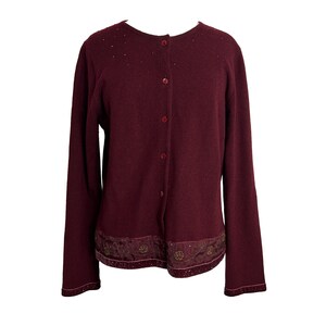 May include: A burgundy cardigan with a button-up front and a decorative trim around the bottom. The trim features a gold floral design and a velvet ribbon.
